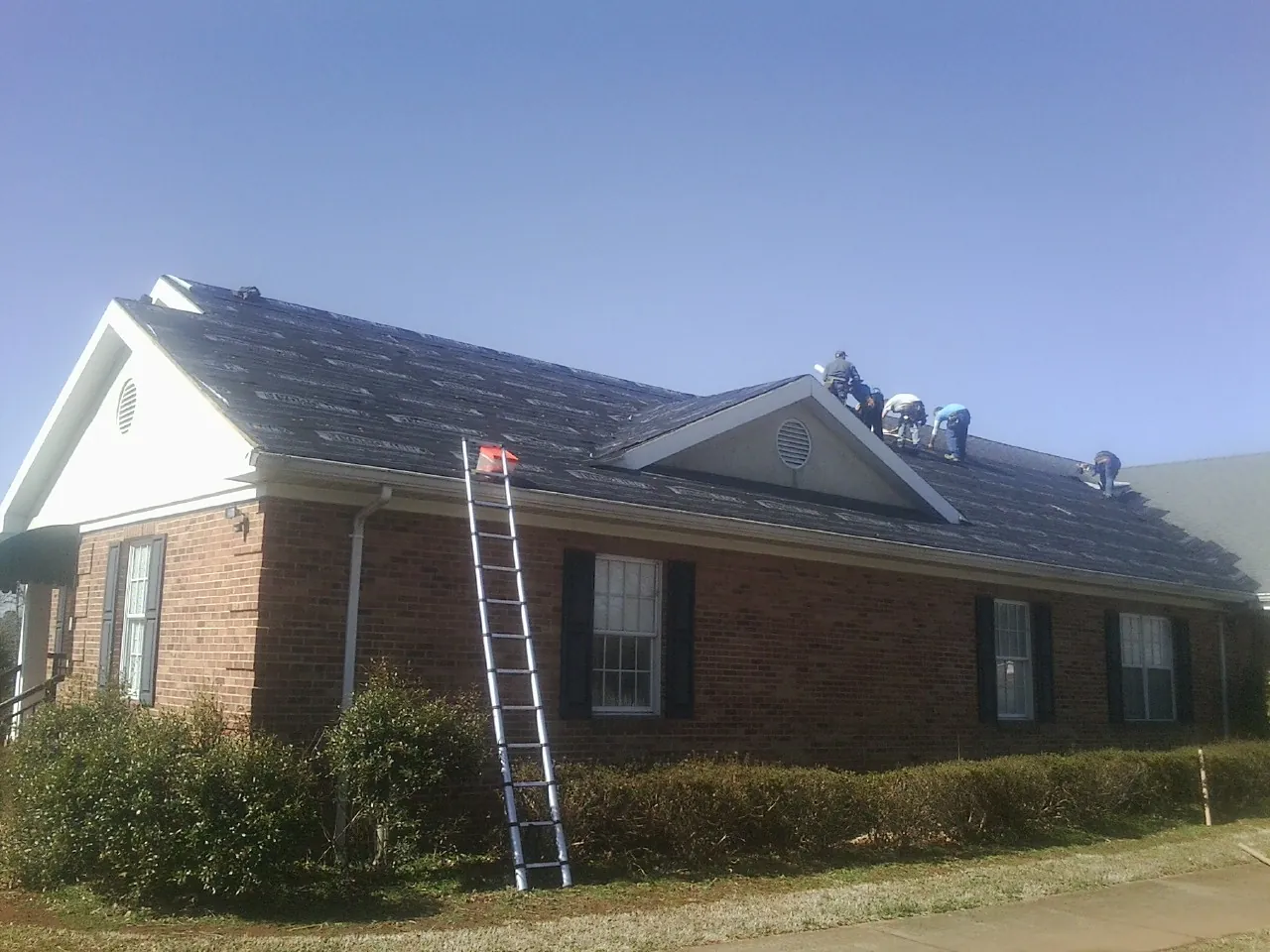 Crew tearing shingles off a brick ranch roof with a ladder set — Emergency Roof Repair work by our crew in Point Lookout