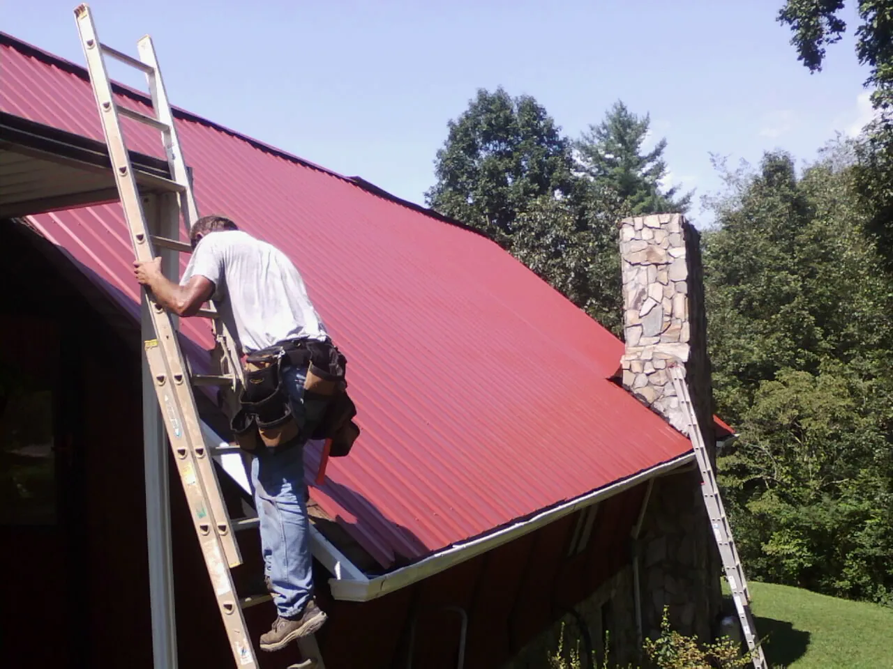 Roofer on a ladder beside a red metal roof and stone chimney — Soffit & Fascia Repair by Roofing Brothers in Point Lookout
